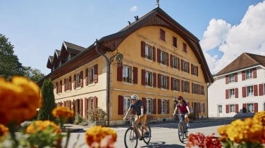 Two people ride bicycles past a house