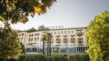 Hotelgebäude mit Wintergarten hinter Bäumen versteckt.