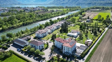 Several white houses next to a river in a green landscape with mountains in the background.