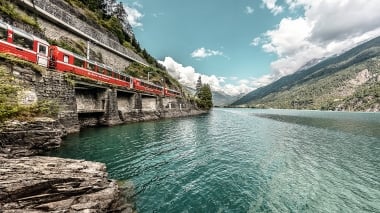 Roter Zug fährt an einer Bergkante entlang eines Sees.