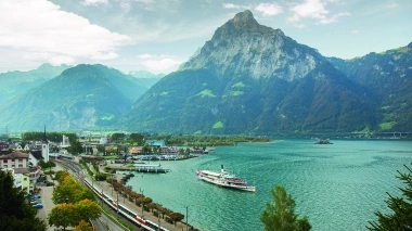 Mountain landscape with lake, train, and ship in the foreground.