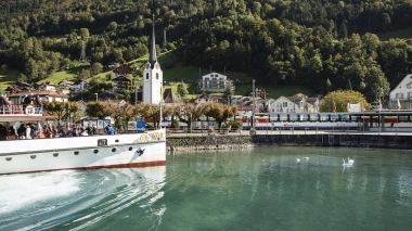Un ferry sur un lac, avec une église et des maisons dans un paysage de montagne verdoyant en arrière-plan.
