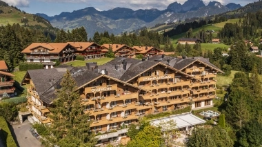 Luftaufnahme des im Chaltstil erbauten Hotels, das von Bäumen umgeben ist und vor einer Bergkulisse liegt.