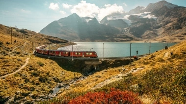 Ein roter Zug der Rhätischen Bahn fährt an einem Bergsee in herbstlicher Stimmung vorbei.