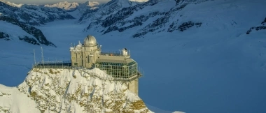 An observatory on a snow-covered mountain peak with mountain landscape in the background.