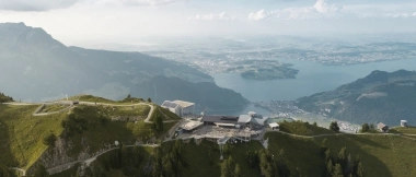 Vista aerea di una cima di montagna con un edificio, circondata da colline verdi e un lago sullo sfondo.