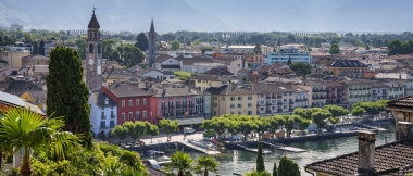 Un panorama cittadino con torri di chiese, edifici colorati e un lago in primo piano, circondato da colline boscose e montagne.