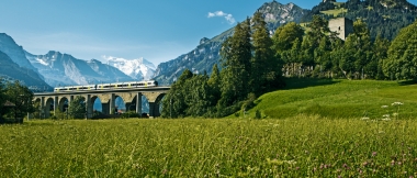 A train crosses a stone bridge in a green mountain landscape.