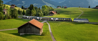 Eine Zugfahrt durch eine grüne Hügellandschaft mit Bergen im Hintergrund.