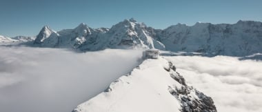 Eine verschneite Berglandschaft mit einem Gebäude auf einem Gipfel über den Wolken.