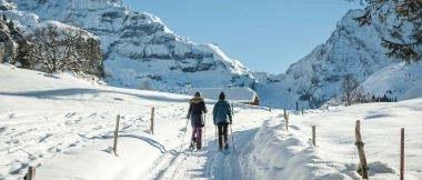 Zwei Personen wandern auf einem verschneiten Weg in einer Winterlandschaft mit Bergen im Hintergrund.