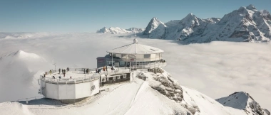 Eine runde Bergstation auf einem verschneiten Gipfel, umgeben von Wolken und schneebedeckten Alpen.