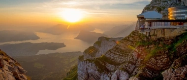 Sonnenuntergang über einer Gebirgslandschaft mit einem Gebäude am Berghang.