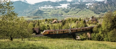 A yellow-orange train travels through a green, hilly landscape with snow-capped mountains in the background.