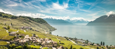 A picturesque landscape with a village, terraced vineyards, and a large lake in the background.