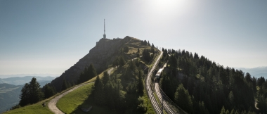 Eine Berglandschaft mit einer Eisenbahnstrecke, die zu einem Gipfel mit einem Sendemast führt, umgeben von Bäumen und Wiesen.