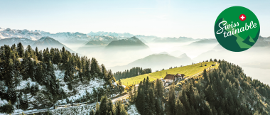 Panoramablick auf die Schweizer Alpen mit Wäldern, Bergen und einem kleinen Bauernhaus, überlagert von einem "Swisstainable"-Logo.