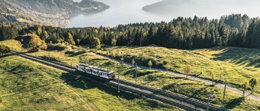 A train travels over green hills with mountains and a lake in the background.