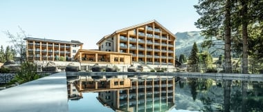 A modern hotel building is reflected in a calm pool, surrounded by mountains and trees.