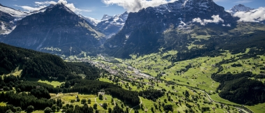 A vast mountain landscape with green meadows, forests, and snow-capped peaks under a blue sky.