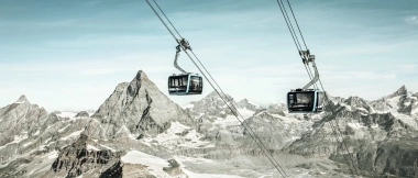 Due cabine della funivia che si muovono davanti a un paesaggio montano alpino.