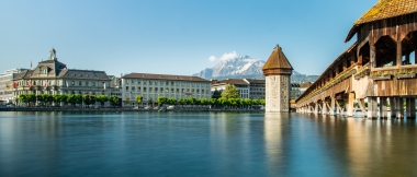 An old wooden bridge and a tower over a calm river, with snow-capped mountains in the background.