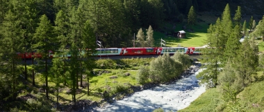 Red train passing through a wooded valley with a river in the foreground.