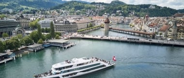 Ein Boot mit Schweizer Flagge fährt auf einem Fluss in einer Stadt mit mittelalterlicher Architektur und einer Holzbrücke, umgeben von grünen Hügeln.
