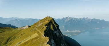 Mountain ridge with a transmission tower, steep slope, and a wide view over a valley with a lake and mountains in the background.