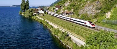 Train traveling along a lake with houses and green hills in the background.