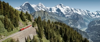 A red train travels through green mountains with snow-covered peaks in the background.