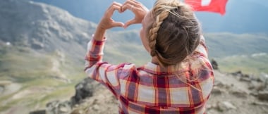 Eine Frau formt ein Herz mit ihren Händen vor einer Schweizer Flagge und Bergkulisse.