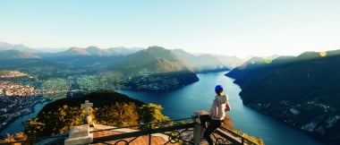 Frau überblickt von einer Terrasse auf einem Berg eine See und Berglandschaft.