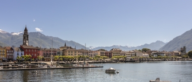 Promenade idyllique au bord du lac avec des maisons colorées et une église en pierre par temps ensoleillé, avec les montagnes en arrière-plan.