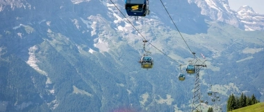 A cable car glides over a green mountain meadow dotted with flowers, set against a stunning mountain backdrop with a view of a valley.