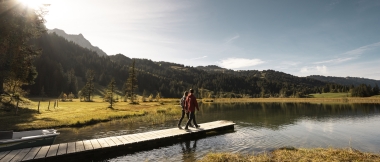Deux personnes marchent sur une passerelle au bord d'un lac entouré d'une forêt aux couleurs d'automne. 