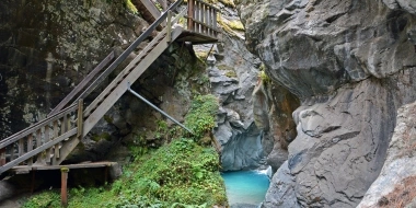 Eine hölzerne Treppe führt durch eine enge, felsige Schlucht mit einem klaren, blauen Wasserbecken am Boden.