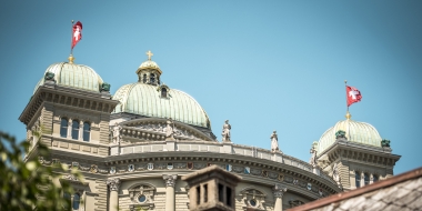 The image shows the Federal Palace in Bern with Swiss flags and a blue sky in the background.