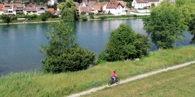 Eine Person fährt auf einem Weg am Fluss entlang; im Hintergrund eine Stadt mit Kirche und grünen Hügeln.