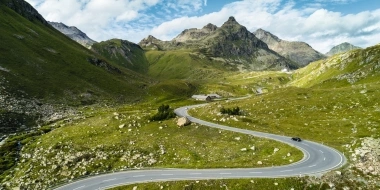 Une route de montagne sinueuse traversant un paysage verdoyant et rocailleux, avec des montagnes en arrière-plan.