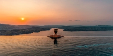 Eine Person entspannt in einem Infinity-Pool mit Blick auf einen See und Sonnenuntergang.