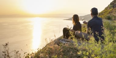 Two people sitting at sunset with a glass of wine on a hillside, looking at the sea.