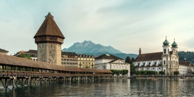 A historic wooden bridge and a tower by the water, with mountains in the background.