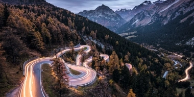 A winding mountain road with light trails at dusk, surrounded by forest and tall mountains in the background.