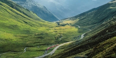 Eine rote Bahn fährt durch ein grünes Alpental mit kurviger Strasse und Bergblick im Hintergrund.