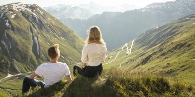 Two people sit on a meadow overlooking a mountainous landscape.