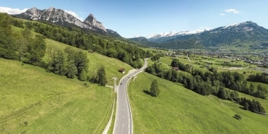 A winding road passes through green hills, surrounded by mountains in the background.