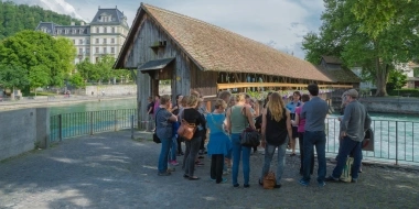 Eine Gruppe von Menschen steht auf einem Platz vor einem überdachten Holzsteg an einem Gewässer, im Hintergrund ein historisches Gebäude und Bäume.