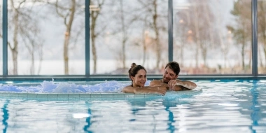 A couple relaxing in the indoor pool with views of trees through large windows.