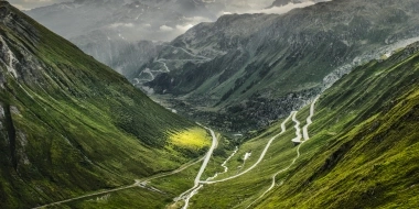 A winding mountain road snakes through green valleys under a dramatic sky.